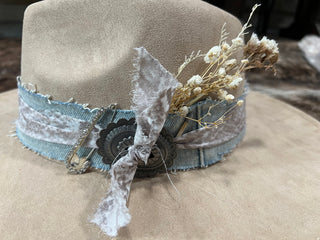Close-up of tan felt flat brim hat band with frayed light-wash denim, antiqued silver concho, rhinestone safety pin and dried flowers