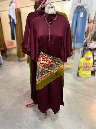Peacock print fringed scarf tied as a hip wrap over a burgundy satin dress with flutter sleeves